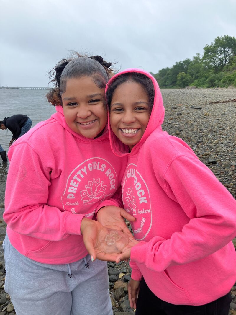 The image shows two young girls smiling and posing for the camera. They are wearing matching pink hoodies that say "PRETTY GIRLS ROCK". They are holding a small, translucent object in their hands, possibly a jellyfish or a similar sea creature. They appear to be at a beach or a shoreline, with water and a distant landmass visible in the background.